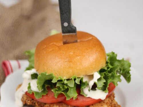 Buttermilk Fried Chicken sandwich on a brioche bun, thick slices of tomato, leaf lettuce, and knife in the center of the bun