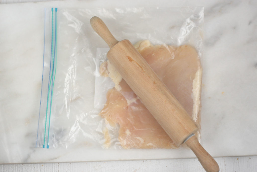 Boneless skinless chicken breast in a large Ziploc bag being flattened with a small wooden rolling pin