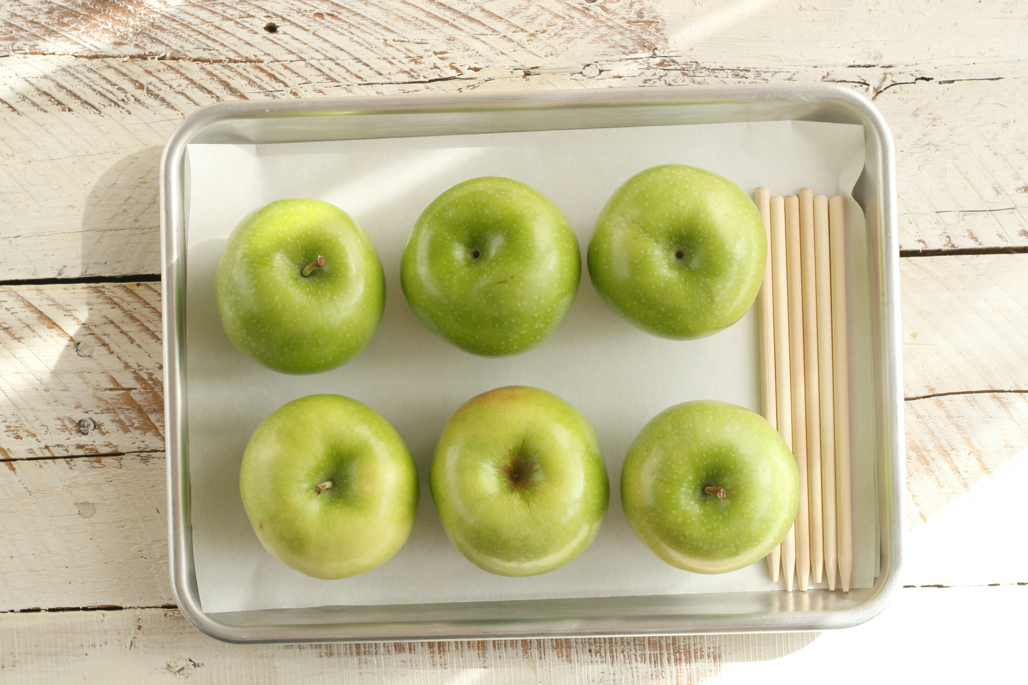 Granny Smith apples with wooden apple sticks sitting on a half sheet pan lined with parchment paper.