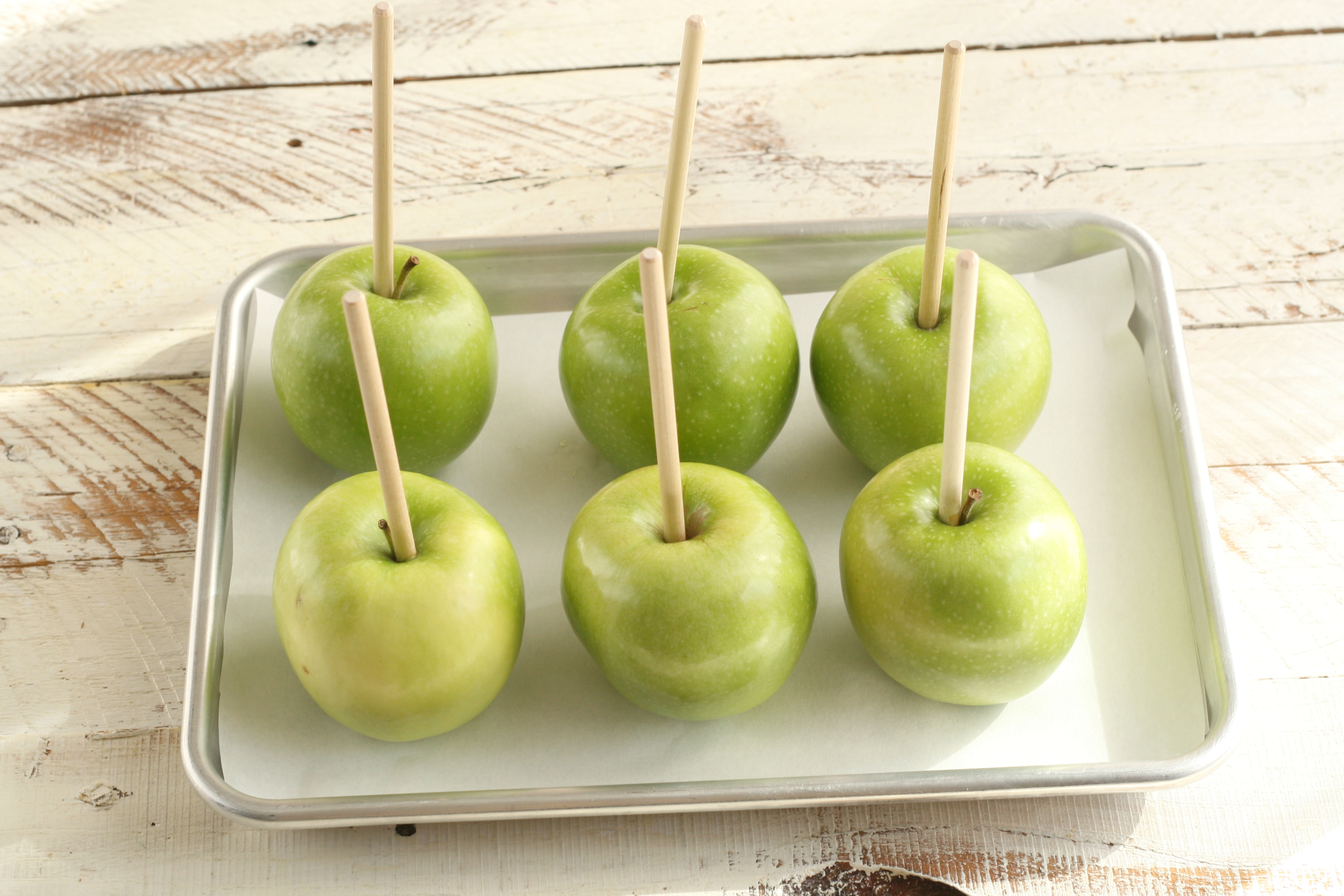 Granny Smith apples with wooden apple sticks sitting on a half sheet pan waiting to be dipped into melted caramel.