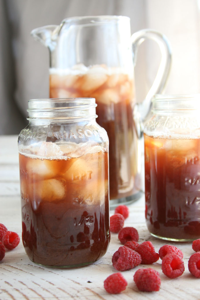 Homemade Raspberry Iced Tea in glass Mason jars, glass pouring pitcher in background, with fresh raspberries sitting around them.