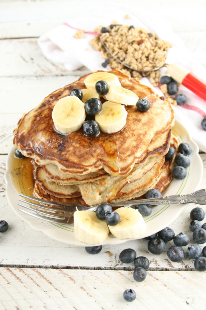 bluberry banana pancakes stacke on plate with maple syrup, fresh banana slices and blueberries