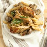 lemon rosemary chicken pasta with a cream sauce and mushrooms on a white plate. Fresh sprig of rosemary on top of pasta