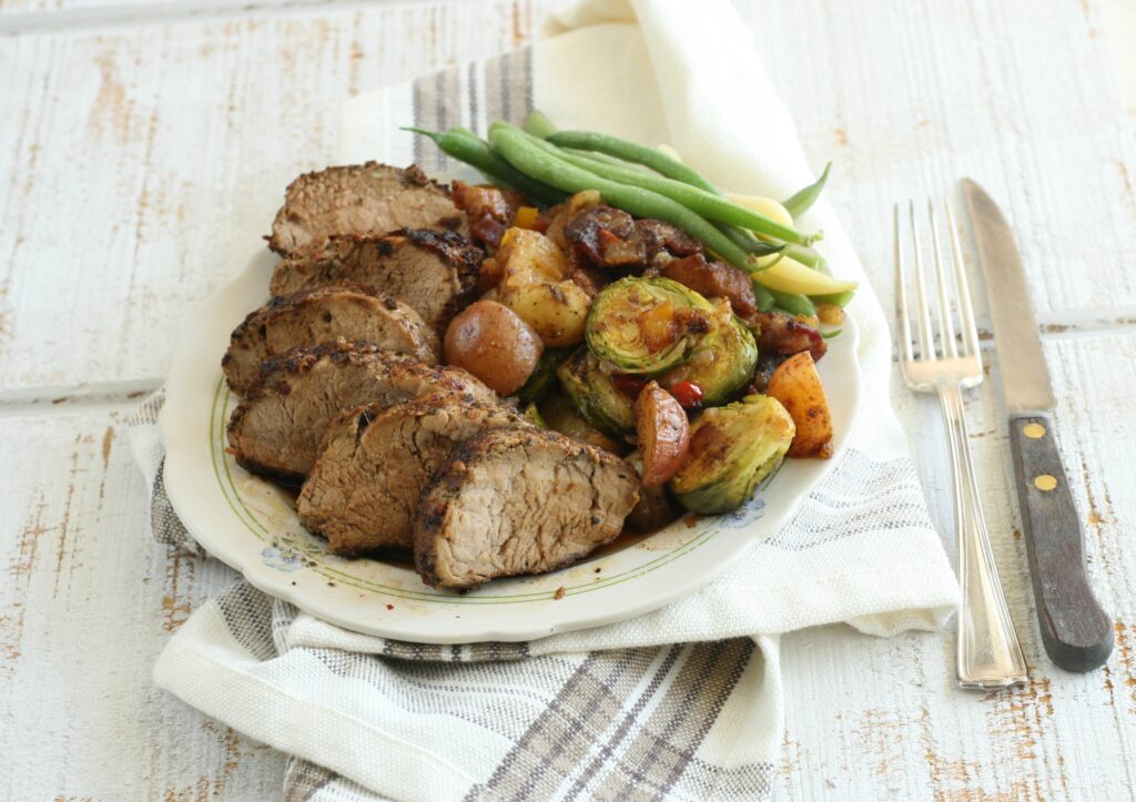 Sliced Pork Tenderloin with Roasted Vegetables on a White Plate with Silverware