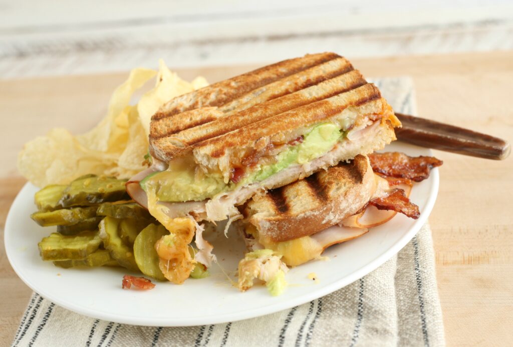 turkey bacon avocado panini on a white plate with potato chips and pickles