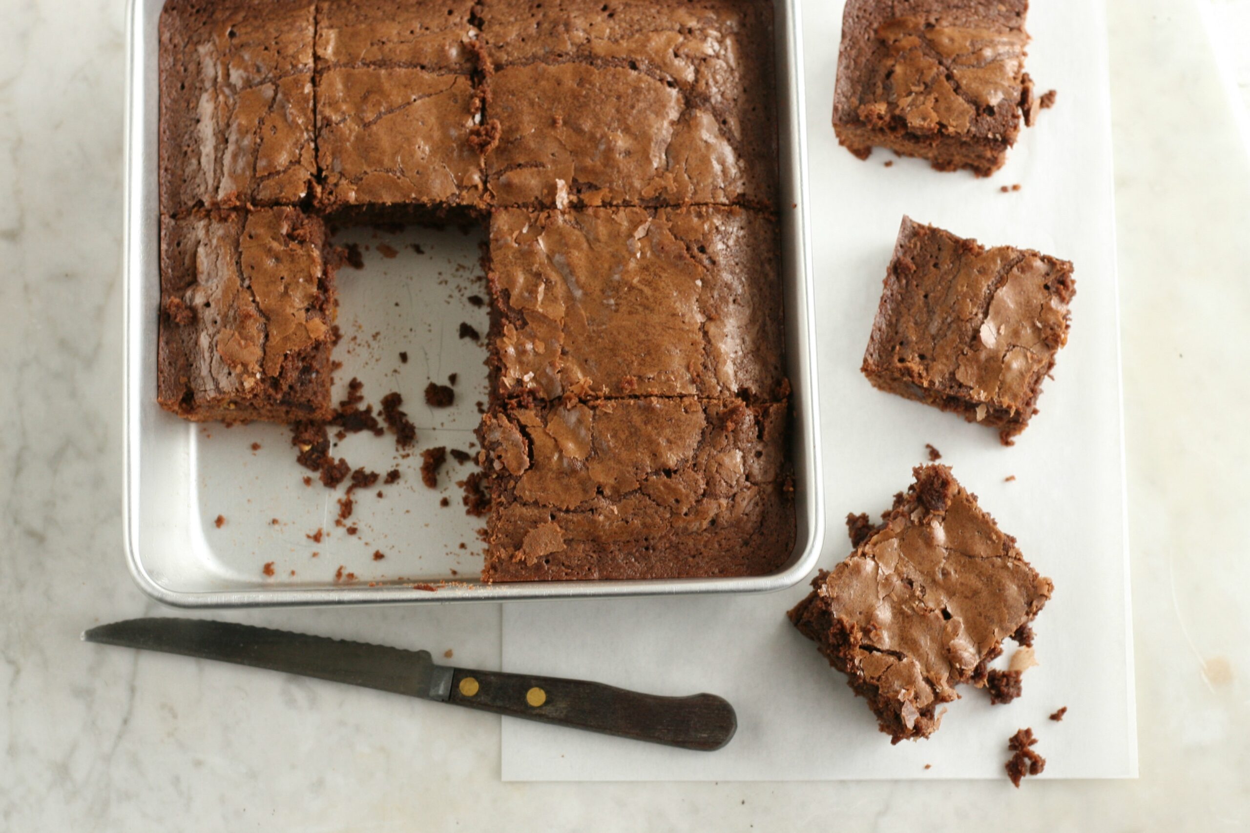 Homemade brownies in square metal baking dish, cut into squares. Some outside the pan to the right.