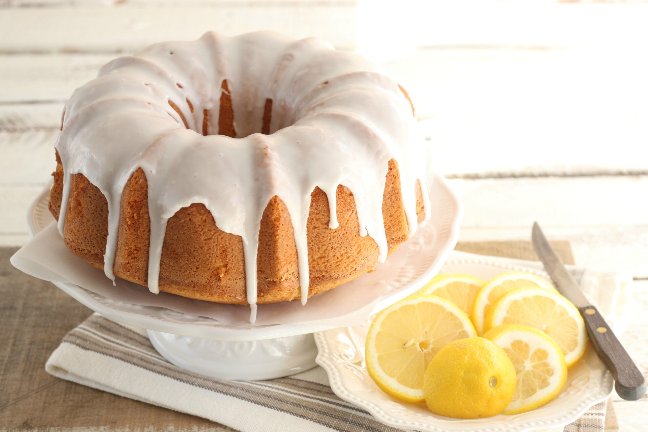 Lemon Bundt cake with icing down sides on white footed cake dish, slices of lemons.