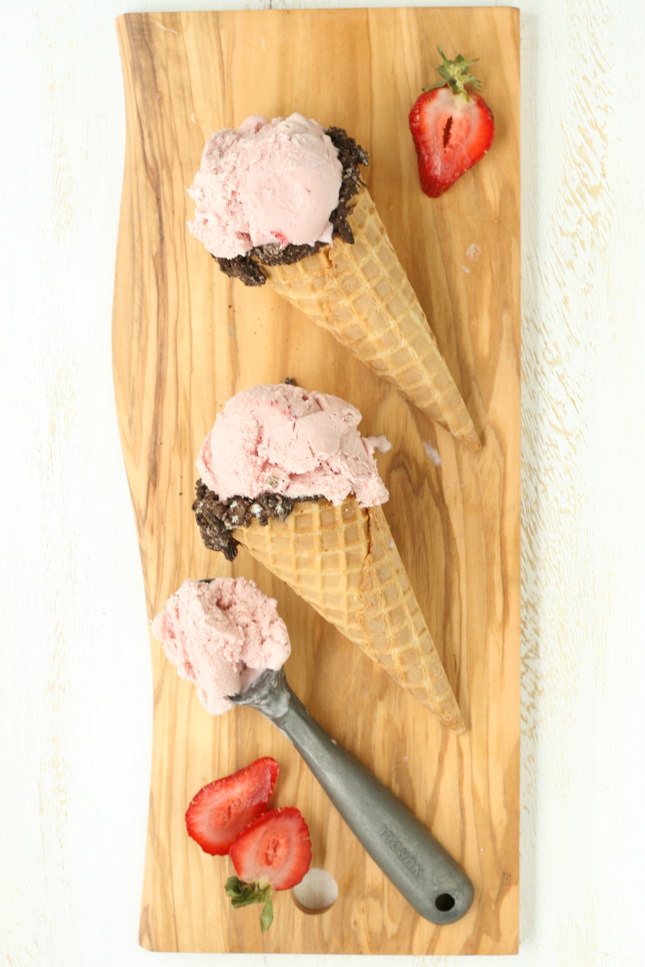 Homemade strawberry ice cream in waffle cones sitting on a wooden cutting board with a vintage ice cream scoop.
