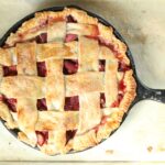 strawberry raspberry rhubarb pie with lattice crust in a cast iron skillet sitting on half sheet pan lined with parchment paper