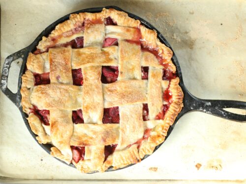 strawberry raspberry rhubarb pie with lattice crust in a cast iron skillet sitting on half sheet pan lined with parchment paper