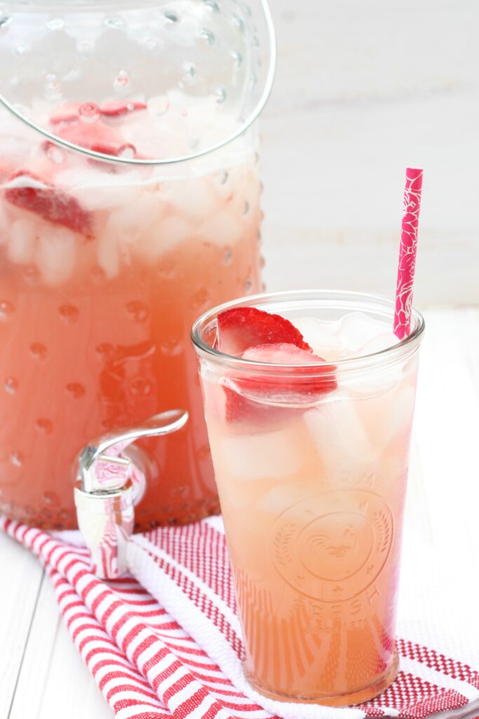 Glass of cold strawberry rhubarb lemonade with raspberry color paper straw and slices of fresh strawberries with ice cubes