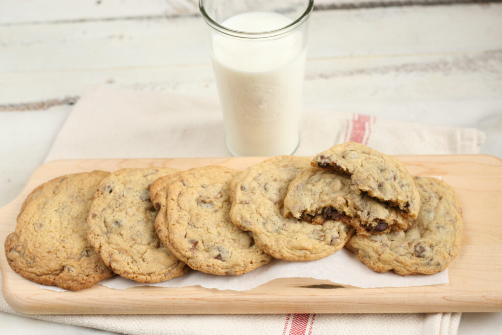 Chocolate chip cookies stacked on each other on wooden cutting board, glass of milk in background