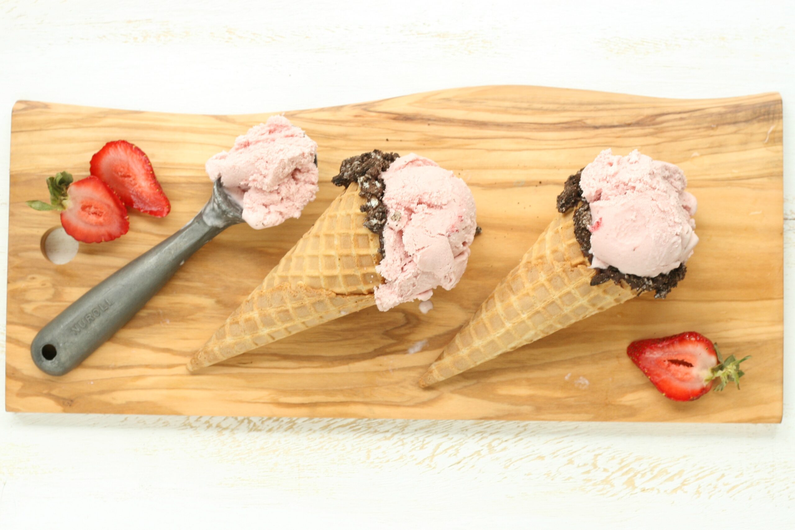 vintage ice cream scoop with strawberry ice cream and two waffle cones dipped in chocolate and crushed Oreo cookies sitting on wooden cutting board.