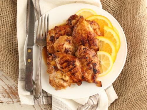 Citrus marinated chicken thighs on a white plate with lemon slices to the right and sitting on white washed vintage wood