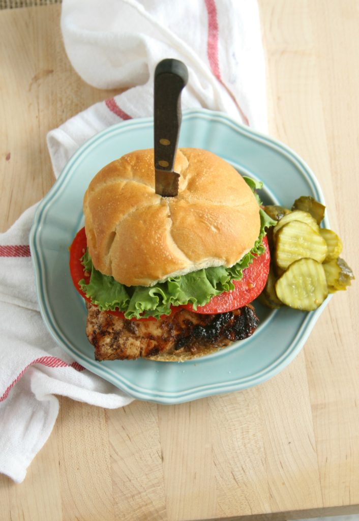 Brown sugar mustard grilled chicken on a crunchy hard roll sitting on a powder blue color plate with slices of heirloom tomatoes, green leaf lettuce, and pickles on the side