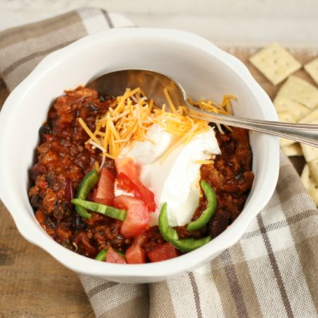 Hearty Chili in a white bowl topped with shredded cheddar cheese, slices of jalapeno peppers, and sour cream