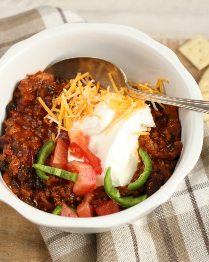 Hearty Chili in a white bowl topped with shredded cheddar cheese, slices of jalapeno peppers, and sour cream