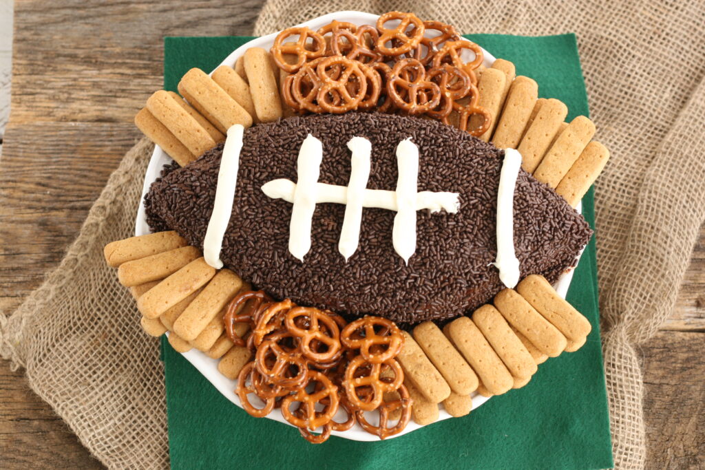 Brownie Batter football dip with chocolate sprinkles and vanilla frosting laces on plate with small pretzels and graham cracker sticks