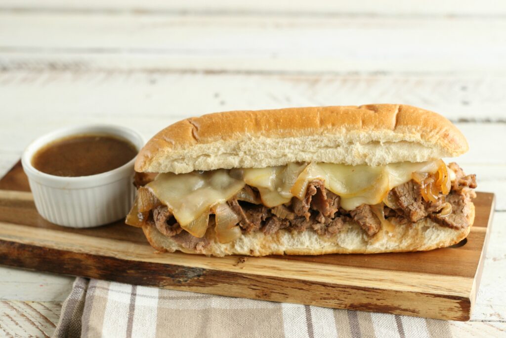 French Dip sandwich with small white crock of au jus sitting on rectangle wooden serving tray.