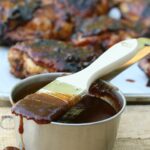 barbecue sauce in metal measuring cup with brush dripping barbecue sauce, chicken in background
