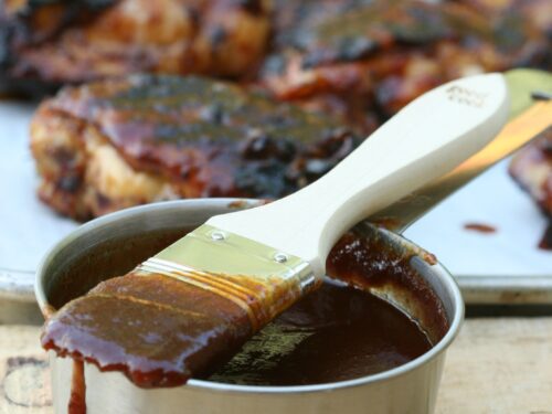 barbecue sauce in metal measuring cup with brush dripping barbecue sauce, chicken in background