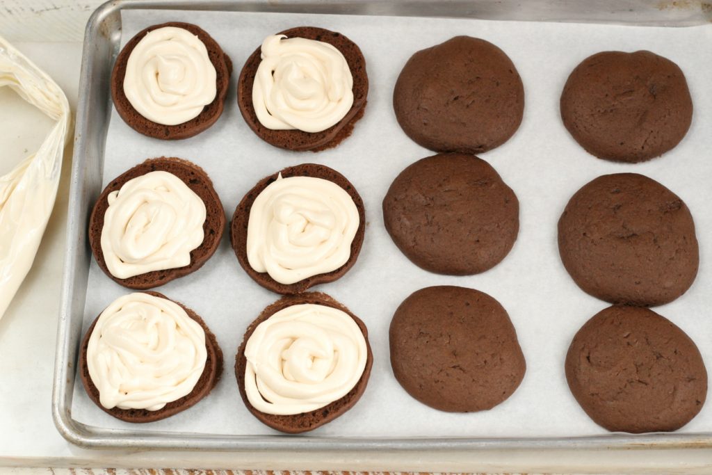 Chocolate Whoopie pies putting frosting on one half on a half sheet pan lined with white parchment paper