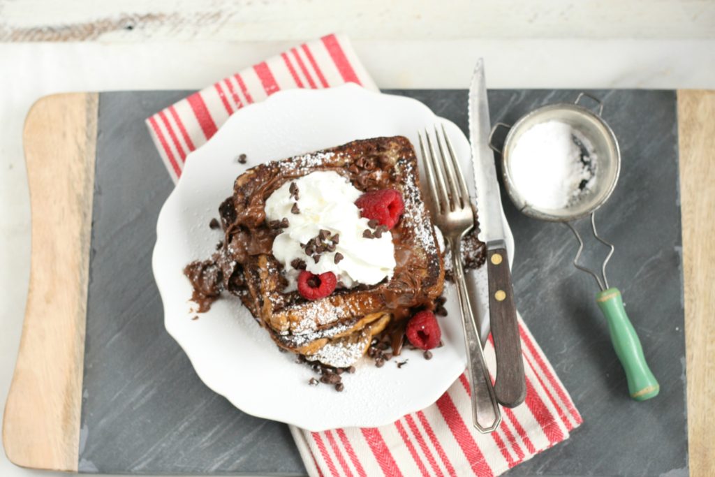 Chocolate Brioche French Toast stacked on a white plate with whipped cream, mini chocolate chips, and fresh raspberries