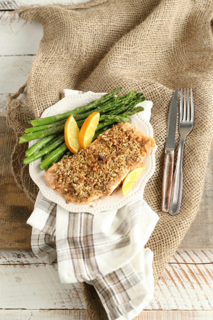 Maple Pecan Crusted Salmon with asparagus and wedges of oranges on white scallop edged plate sitting on a piece of burlap