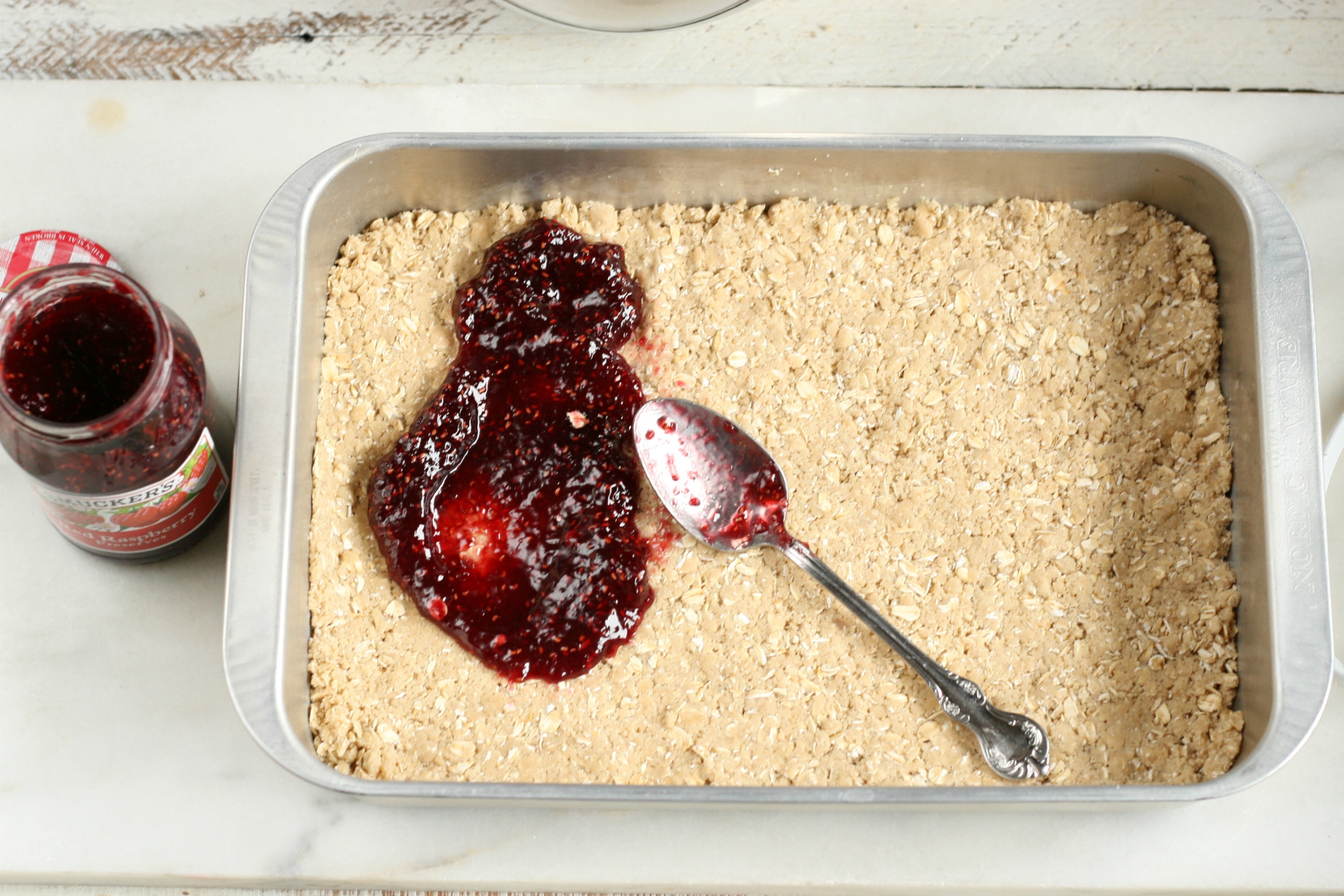 Raspberry Oatmeal Bars pressed into a baking pan and spreading raspberry jam layer with a spoon.