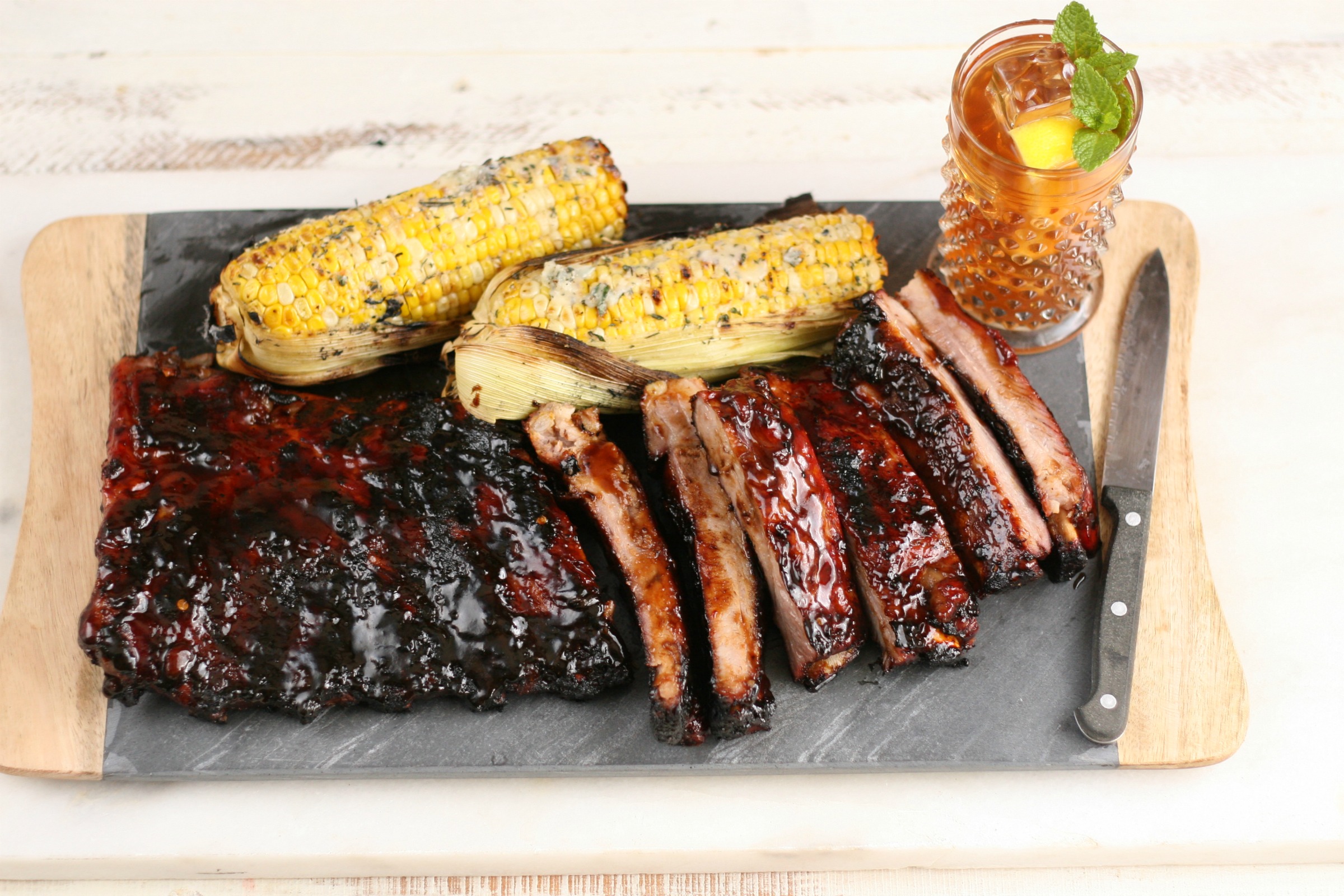 BBQ Ribs on gray slate tray sliced with small knife and two pieces of grilled corn.