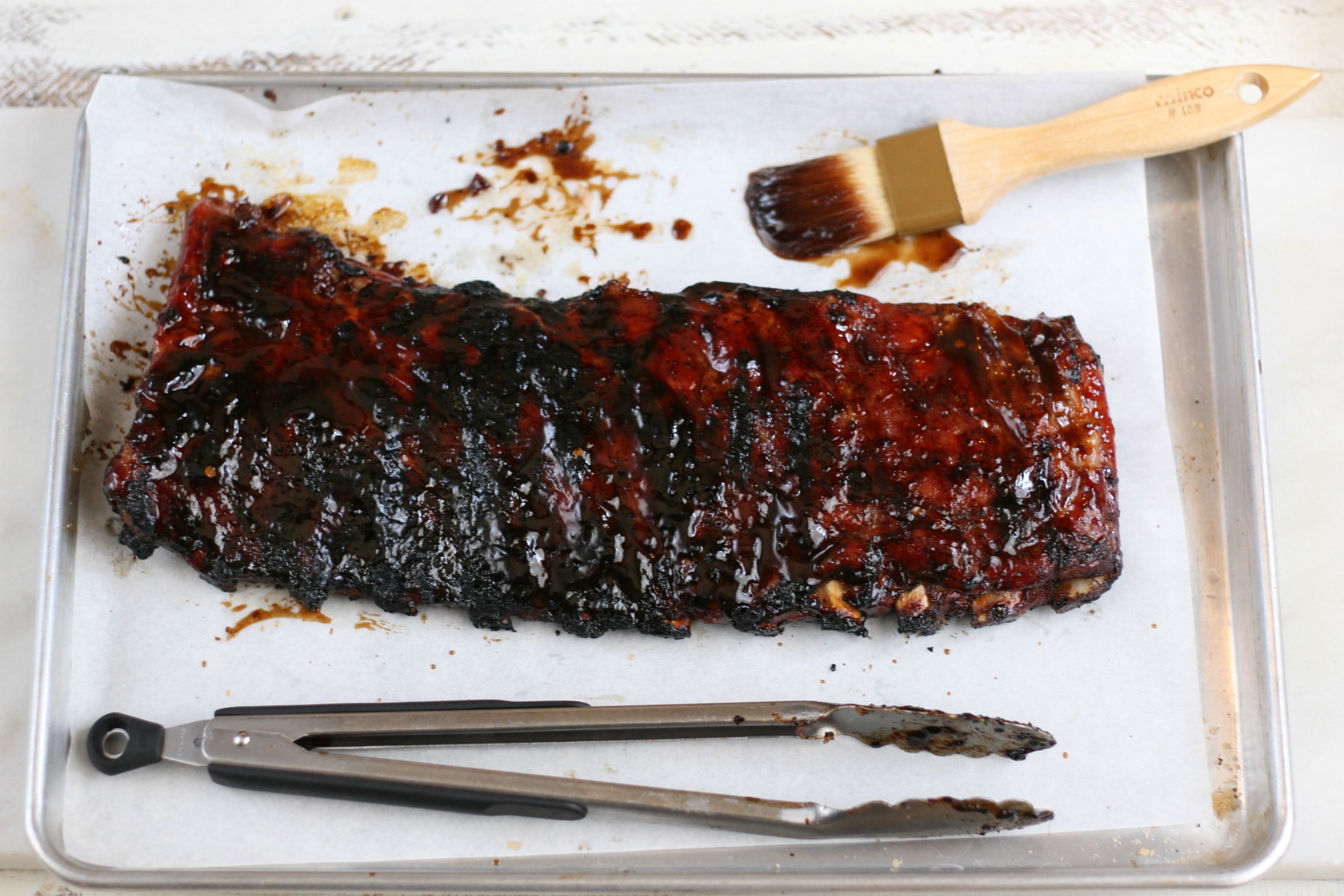 Ribs sitting on half sheet pan, basting brush with barbecue sauce and pair of tongs.