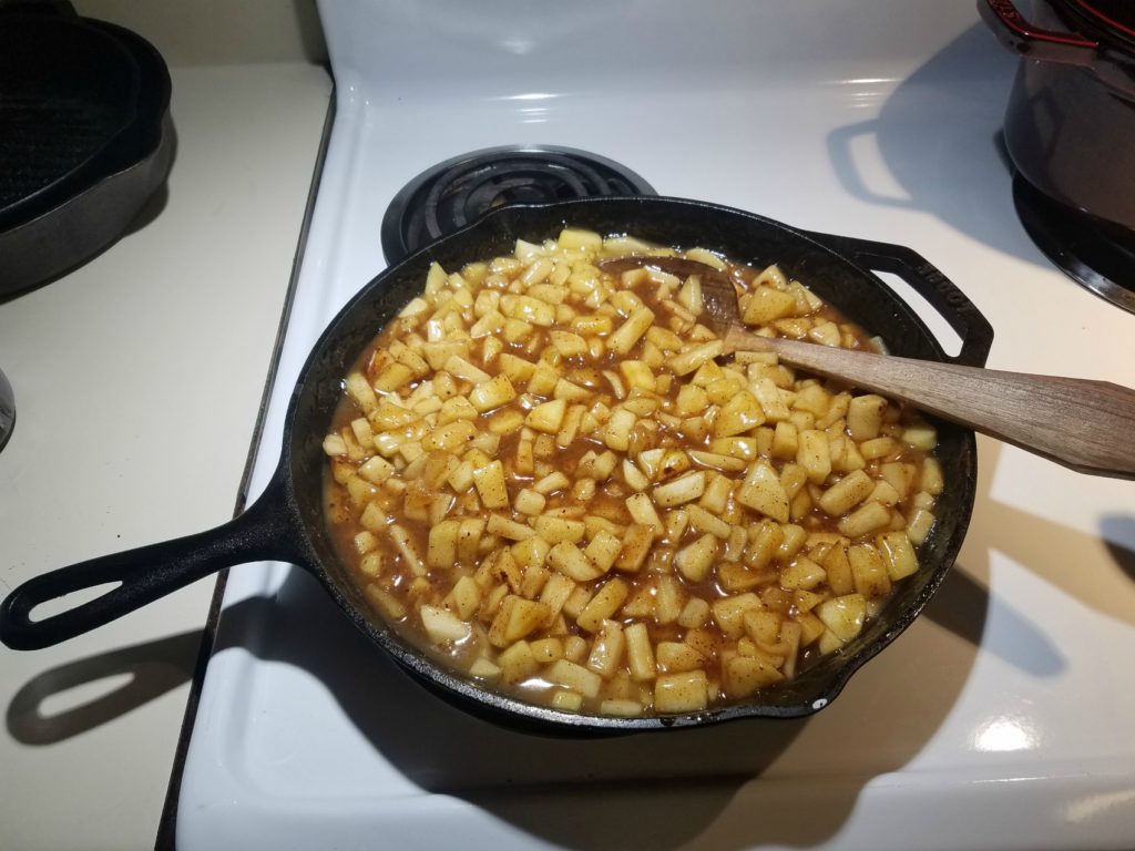 apple pie filling in a large cast iron skillet with wooden spoon on an white kitchen stove.
