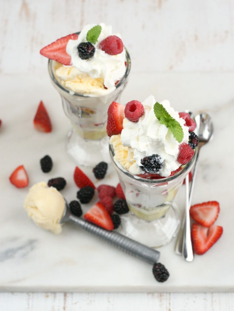 Strawberry Trifle in glass Sundae cups, topped with whipped cream, fresh mint leaf, scoop of vanilla ice cream on white marble