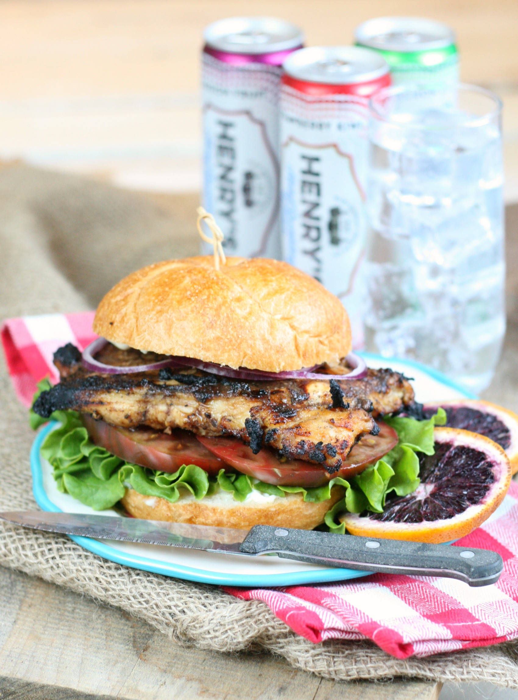 Grilled Chicken sitting on roll with tomatoes, lettuce, and thin slices of red onion, slices of blood oranges on the side of the plate.