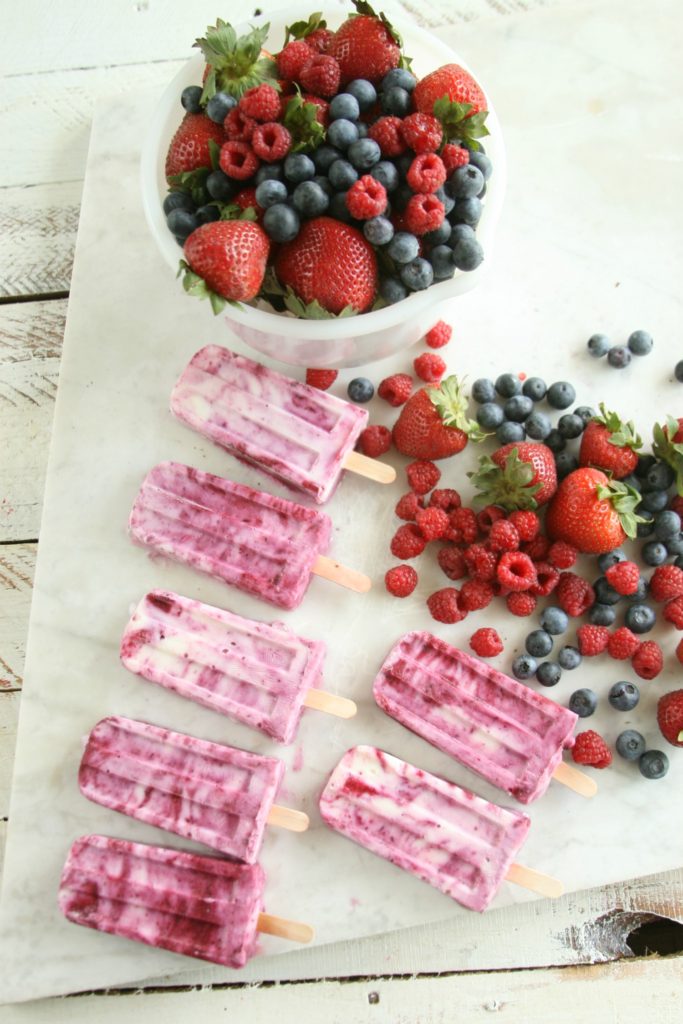 Berry Swirl Yogurt pops lined up on a piece of white marble with a vintage white ceramic bowl of fresh raspberries, strawberries, and blueberries