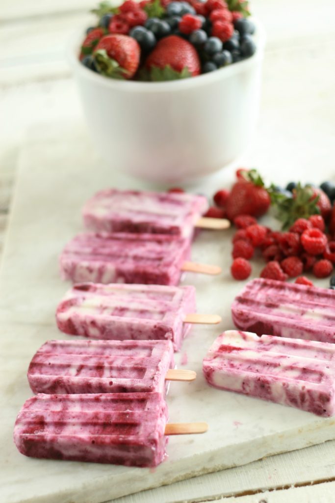 Berry Swirl Yogurt pops lined up with a bowl of fresh raspberries, strawberries, and blueberries in the background