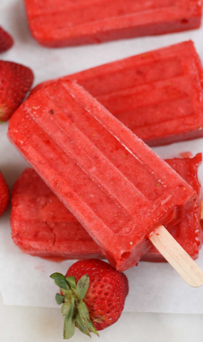strawberry popsicles with wooden sticks stacked on each other on white marble, a few fresh strawberries scattered around.