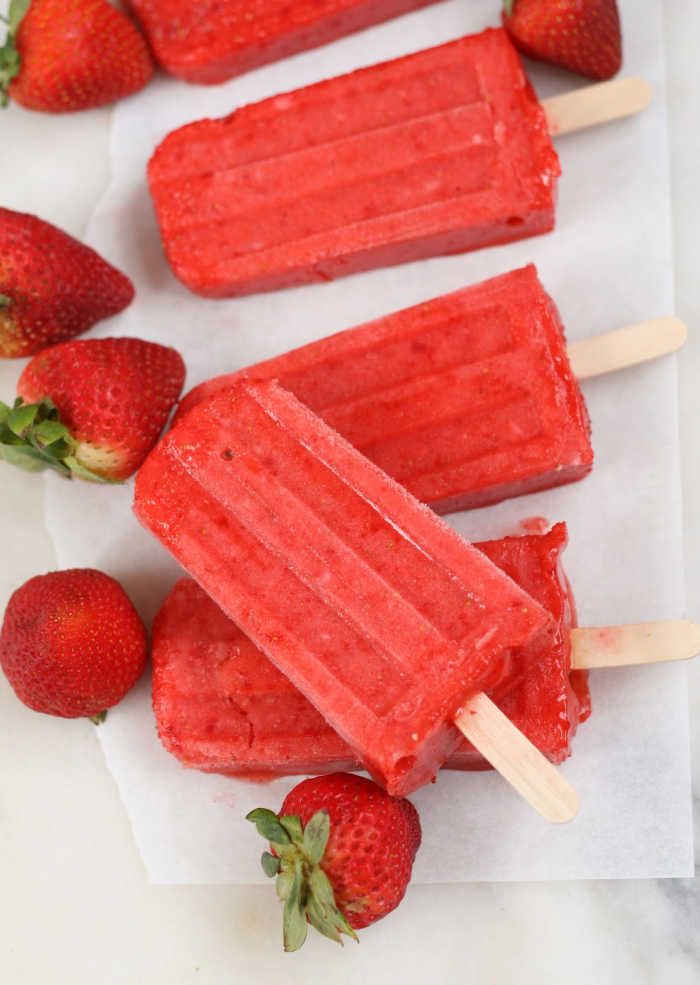 strawberry popsicles sitting on each other on white marble, fresh strawberries scattered around them.
