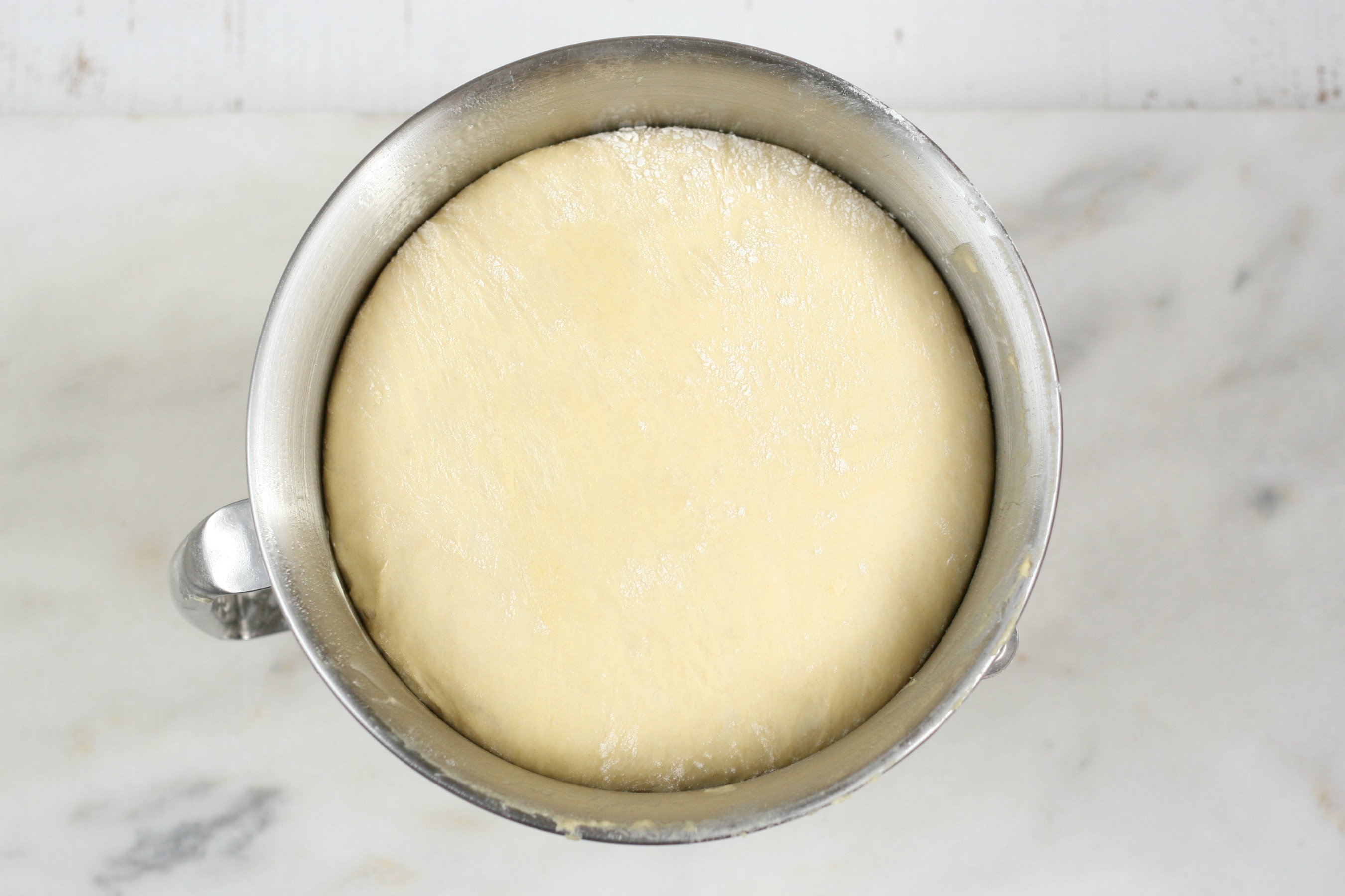 pizza dough rising in a metal mixing bowl on white marble counter.