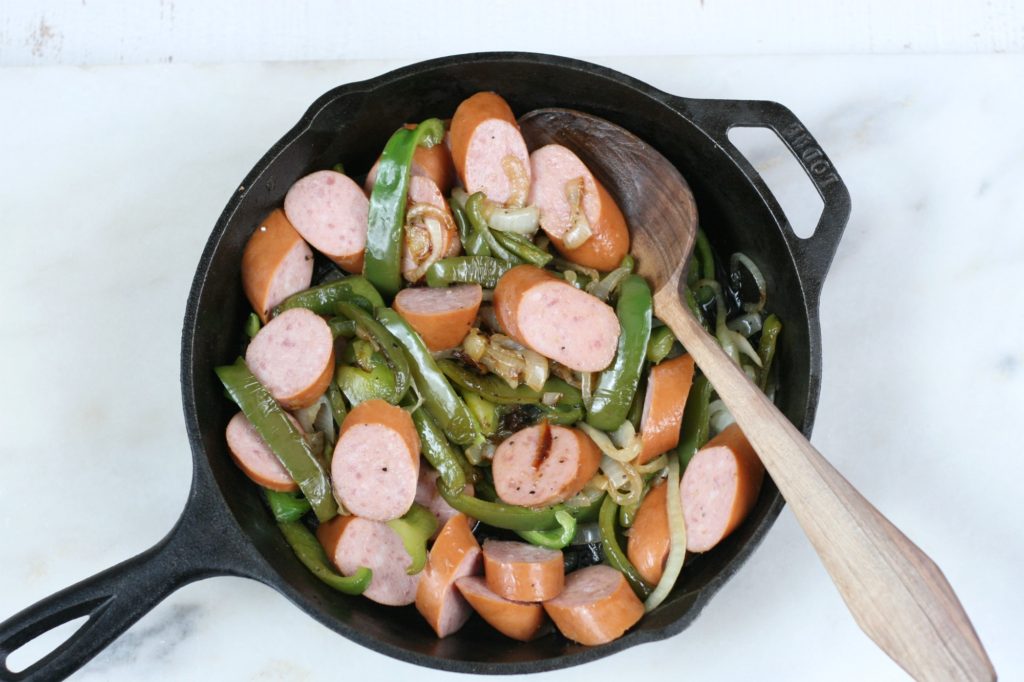 cooked smoked sausage with green peppers and onions in a small cast iron skillet with a handmade wooden spoon