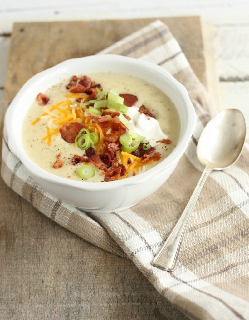 loaded baked potato soup in a white ceramic bowl topped with crumbled bacon, sour cream, shredded cheese, and green onion slices