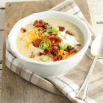 loaded baked potato soup with crumbled bacon pieces, shredded cheddar cheese, sour cream, and thin slices of green onions in a white glass bowl with large spoon to the right of the bowl