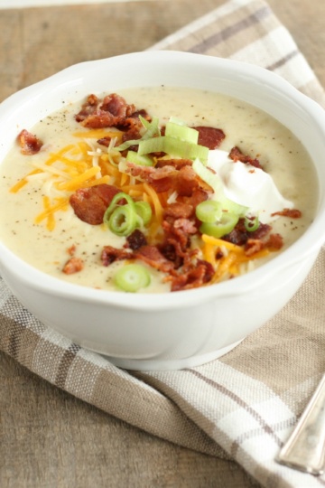 loaded baked potato soup with crumbled bacon pieces, shredded cheddar cheese, sour cream, and thin slices of green onions in a white glass bowl with large spoon to the right of the bowl
