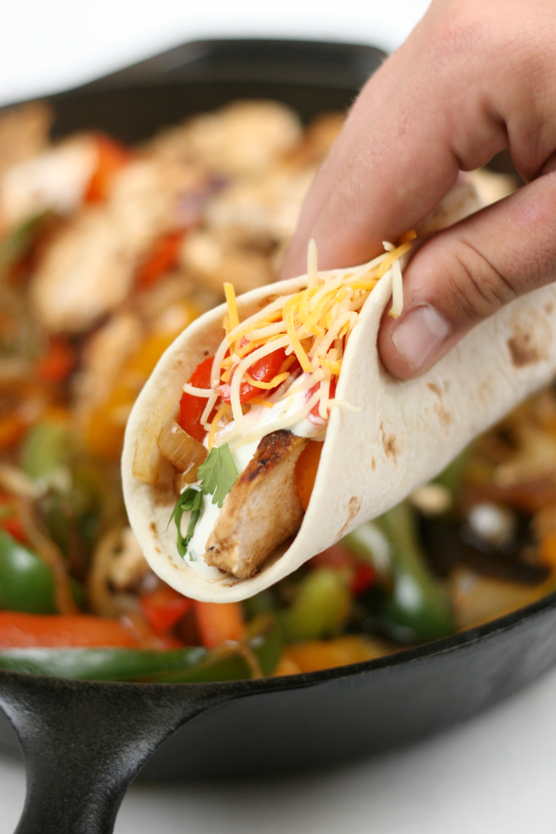 hand holding a chicken fajita with cast iron skillet with onions, peppers, and grilled chicken in the background.