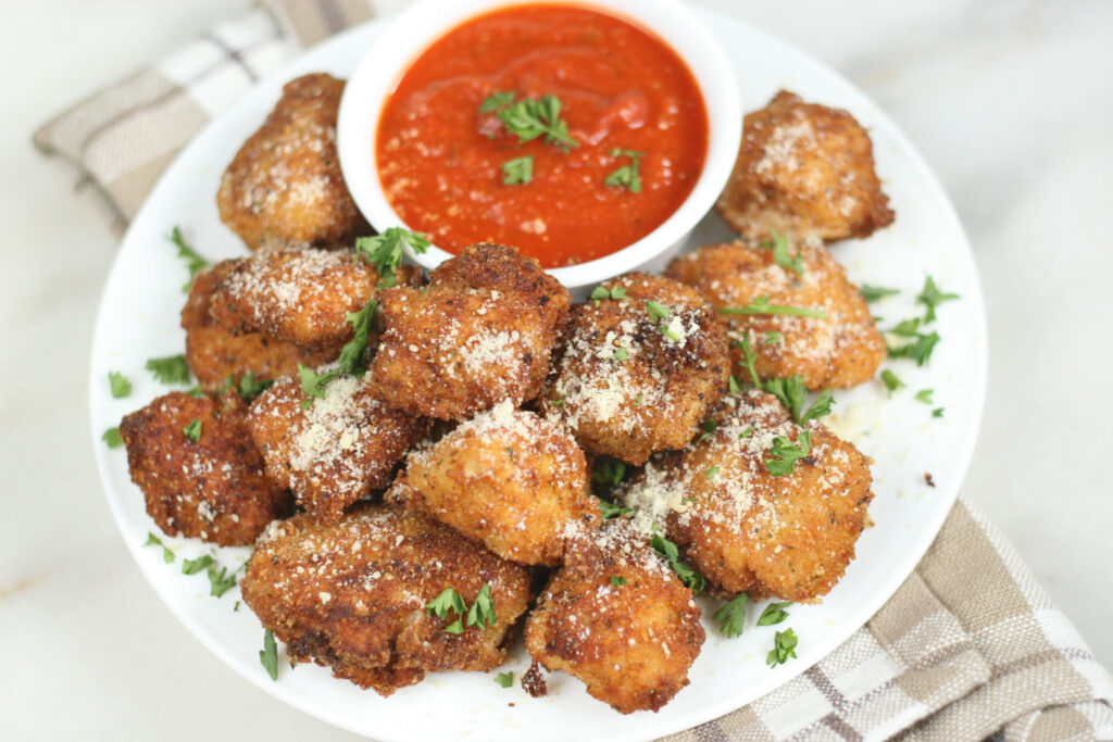 parmesan chicken bites piled up high on a small plate and served with a small cup of warm marinara sauce