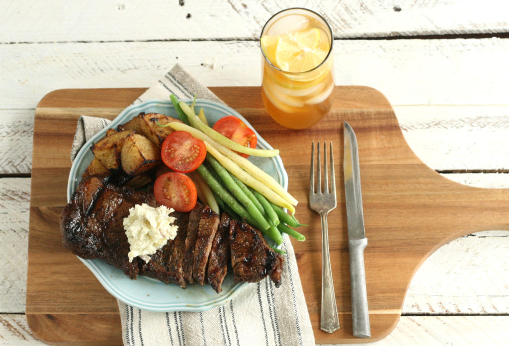 Whiskey Marinated Steak on a cutting board with sliced tomatoes, grilled potatoes, and freshly picked green beans, sitting on a plate on top of a wooden cutting board with handle