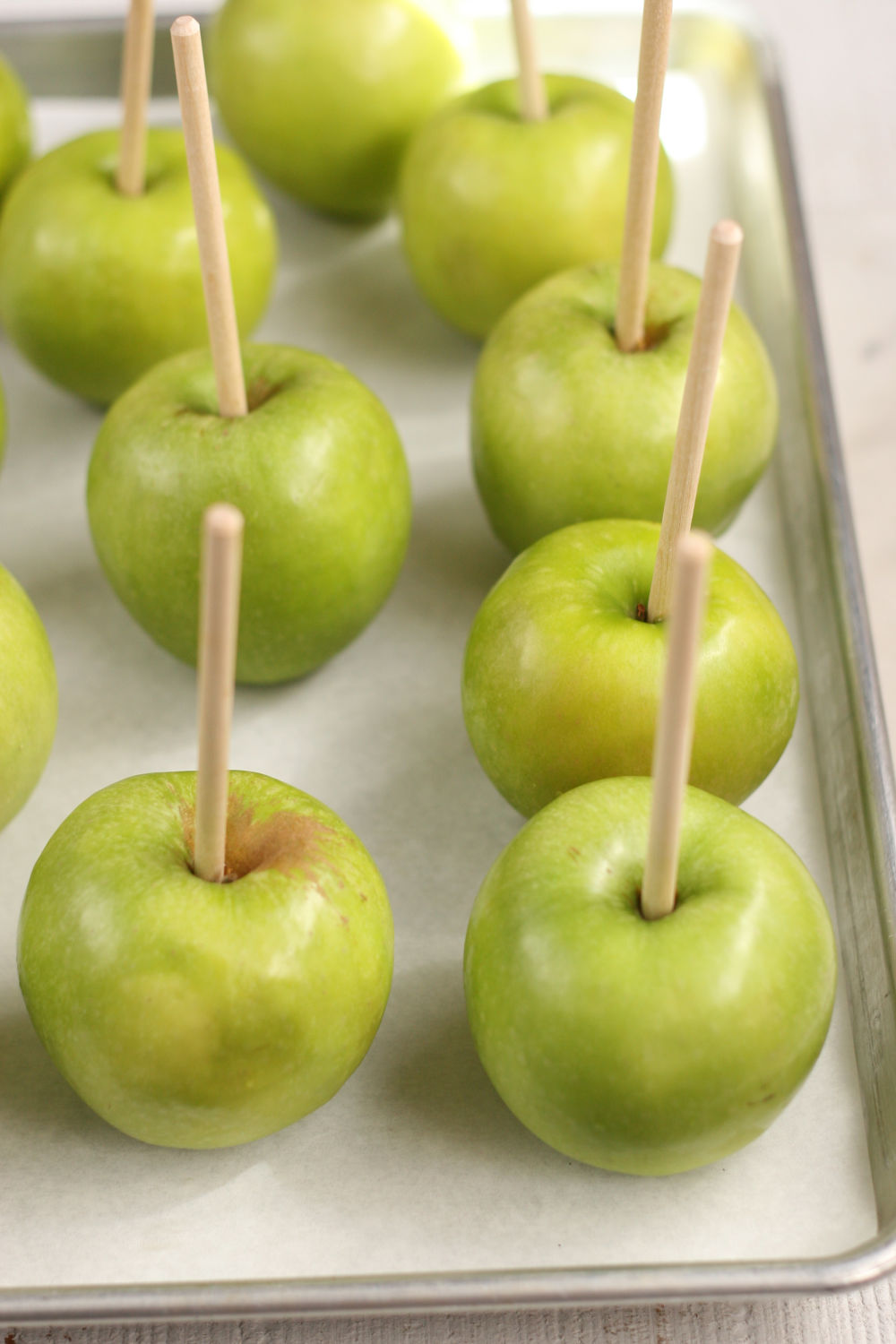 Granny Smith apples with wooden sticks inserted on a half sheet pan lined with parchment paper.