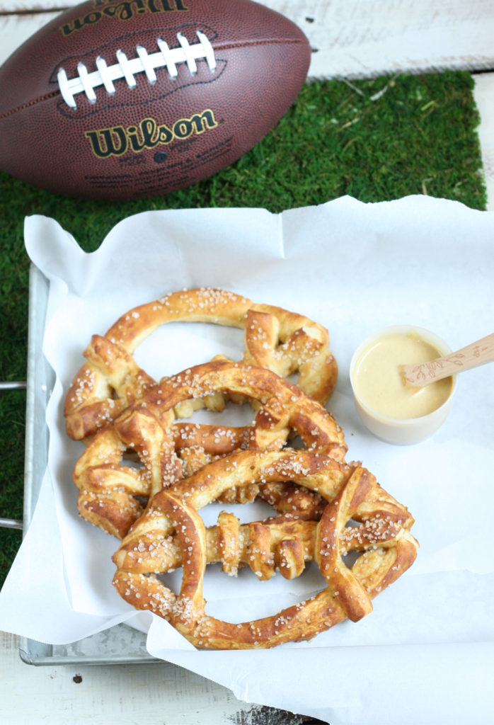 Salted Football pretzels sitting on a galvanized tray with a small bowl of honey mustard and a real football in the background