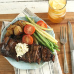 whiskey marinated steak sitting on a dish with fresh tomato slices, green beans, yellow wax beans and roasted potatoes