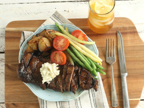 whiskey marinated steak sitting on a dish with fresh tomato slices, green beans, yellow wax beans and roasted potatoes